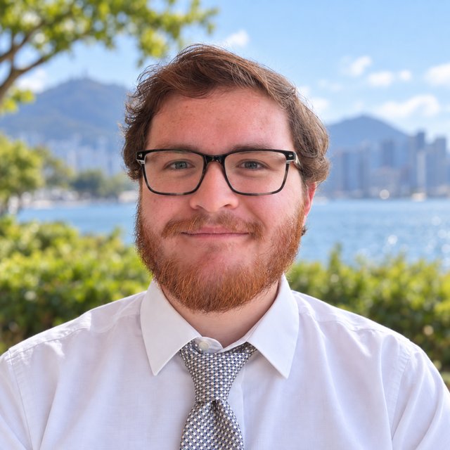 Portrait of Daniel Flippo: a smiling man with auburn hair, a red beard, dark-framed glasses, a white shirt and a patterned tie, with a harbor and hills blurred behind him.